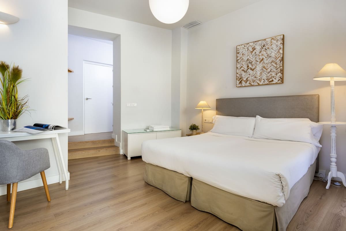 A modern hotel room with a large bed, a desk with a chair, a bedside table with a lamp, and a door leading to another room.
