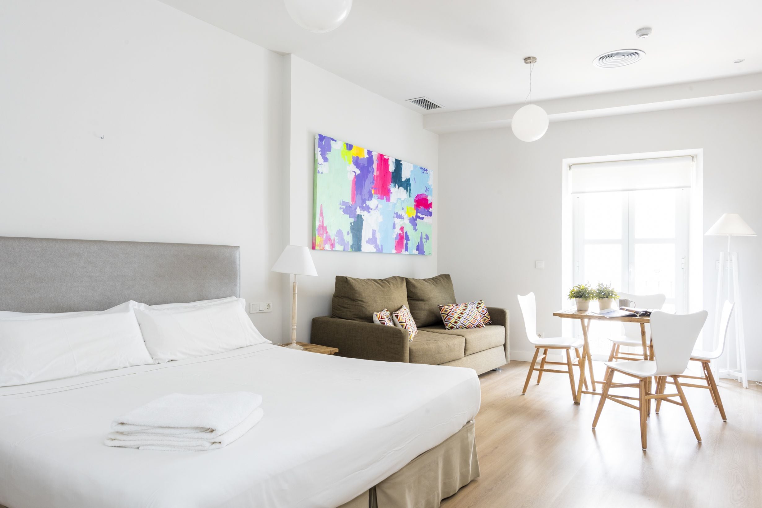 A modern hotel room with a large bed, a sofa, a dining table with chairs, and a colorful abstract painting on the wall.