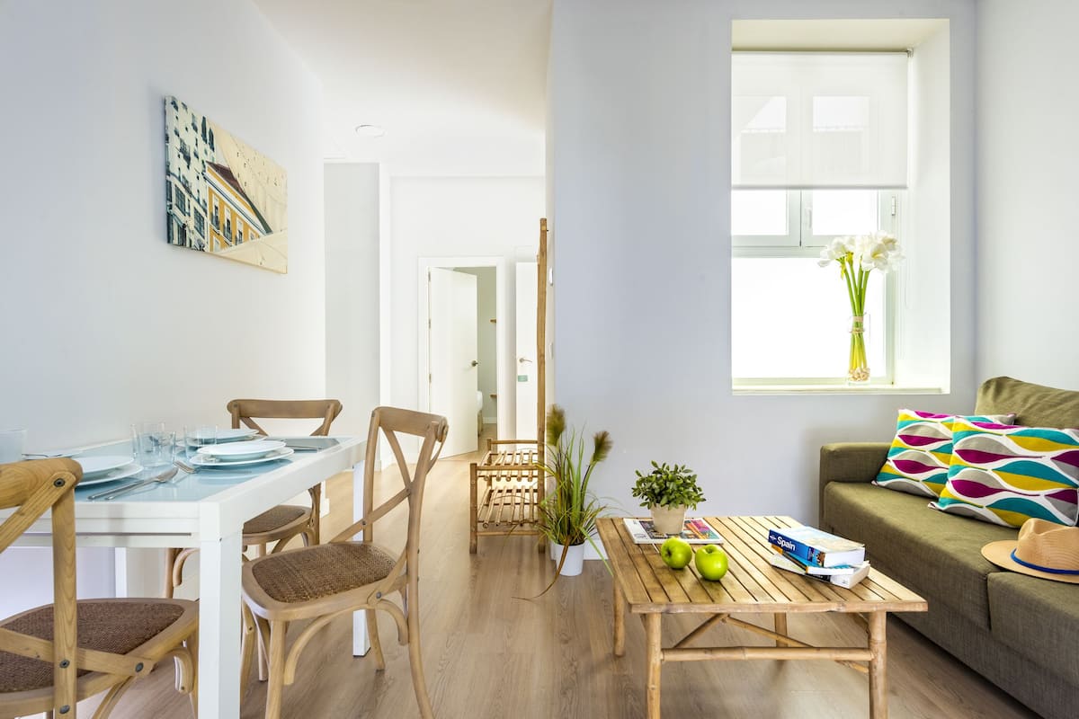 A modern living room with a dining area, a sofa, a window with a view, and a table with books and apples.