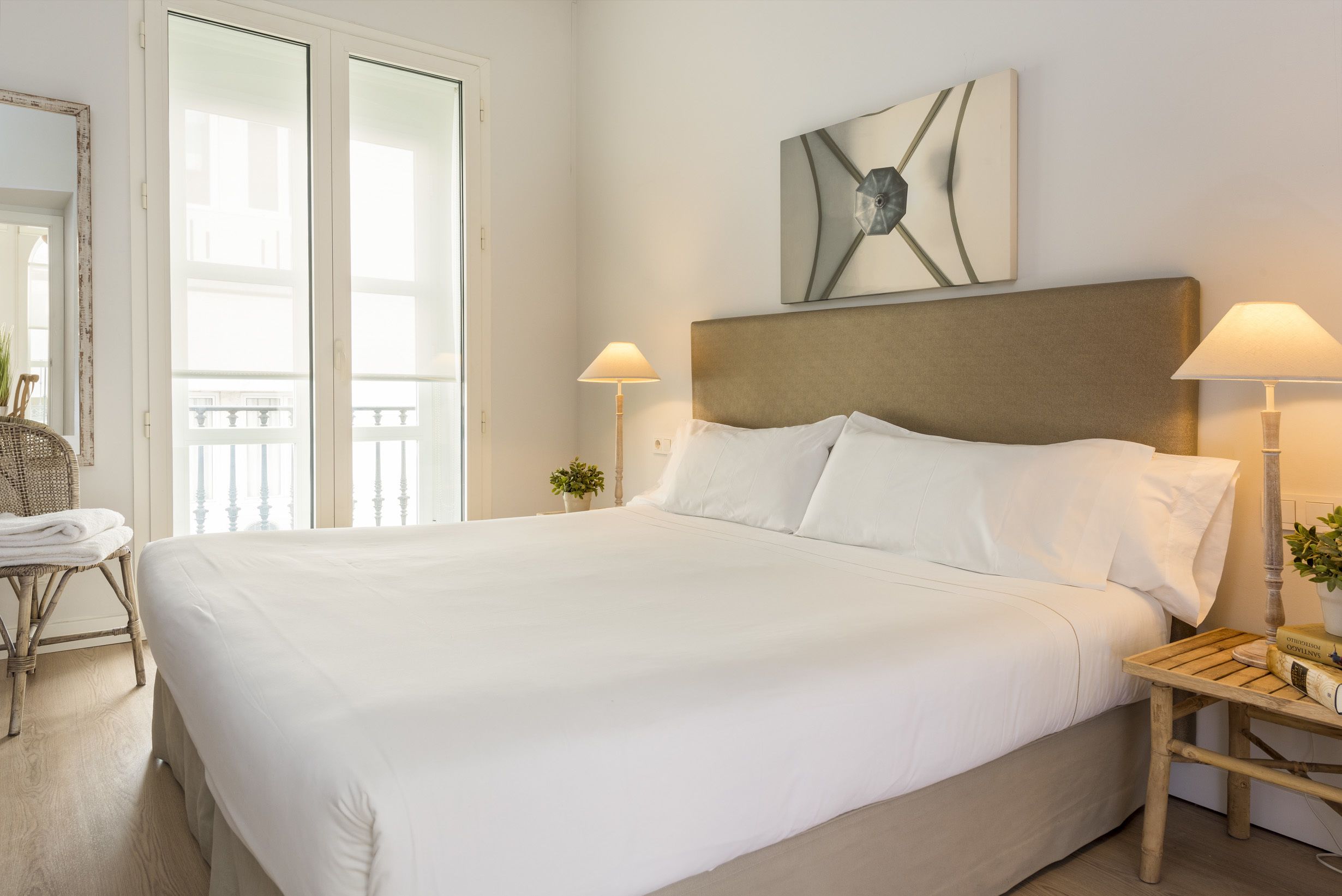 Una cama grande con ropa blanca, dos lámparas de noche, un mesita de noche de madera y una obra de arte abstracta colgada en la pared.