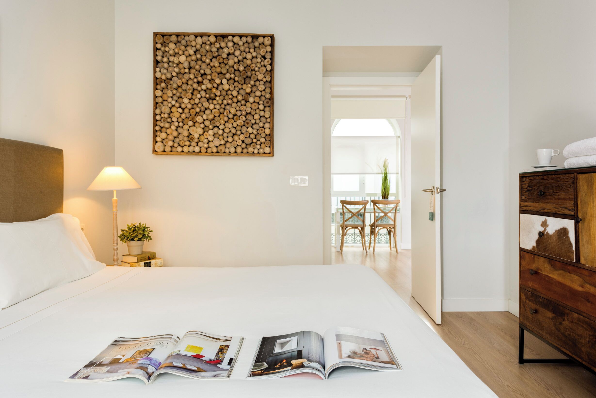 Une chambre d’hôtel avec un lit, un magazine posé sur le lit, une lampe, une tête de lit en bois, une armoire en bois et une vue sur le balcon.