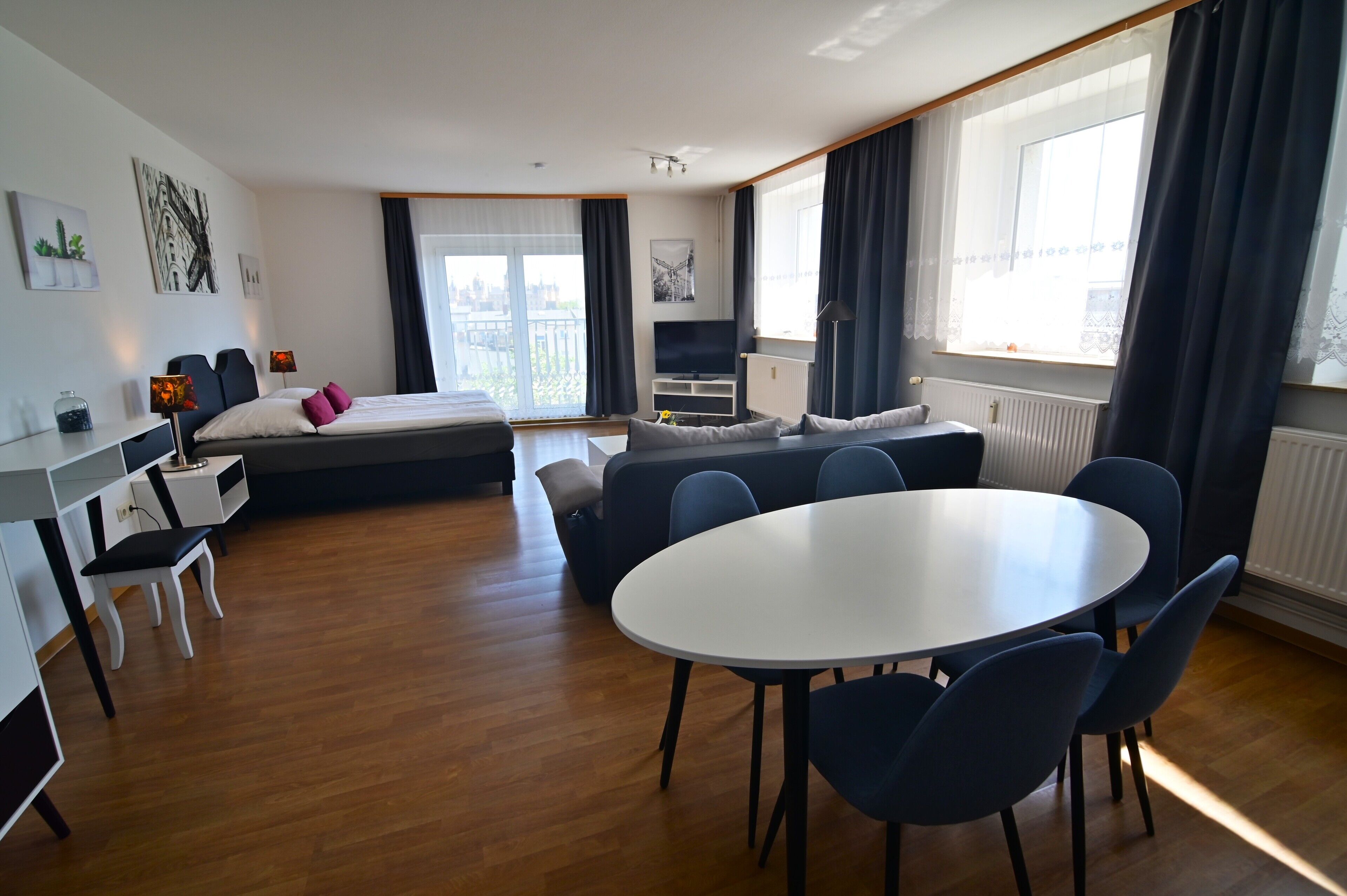 Una habitación de hotel moderna con cama, sofá, mesa de comedor con sillas, televisor y un amplio ventanal con cortinas.