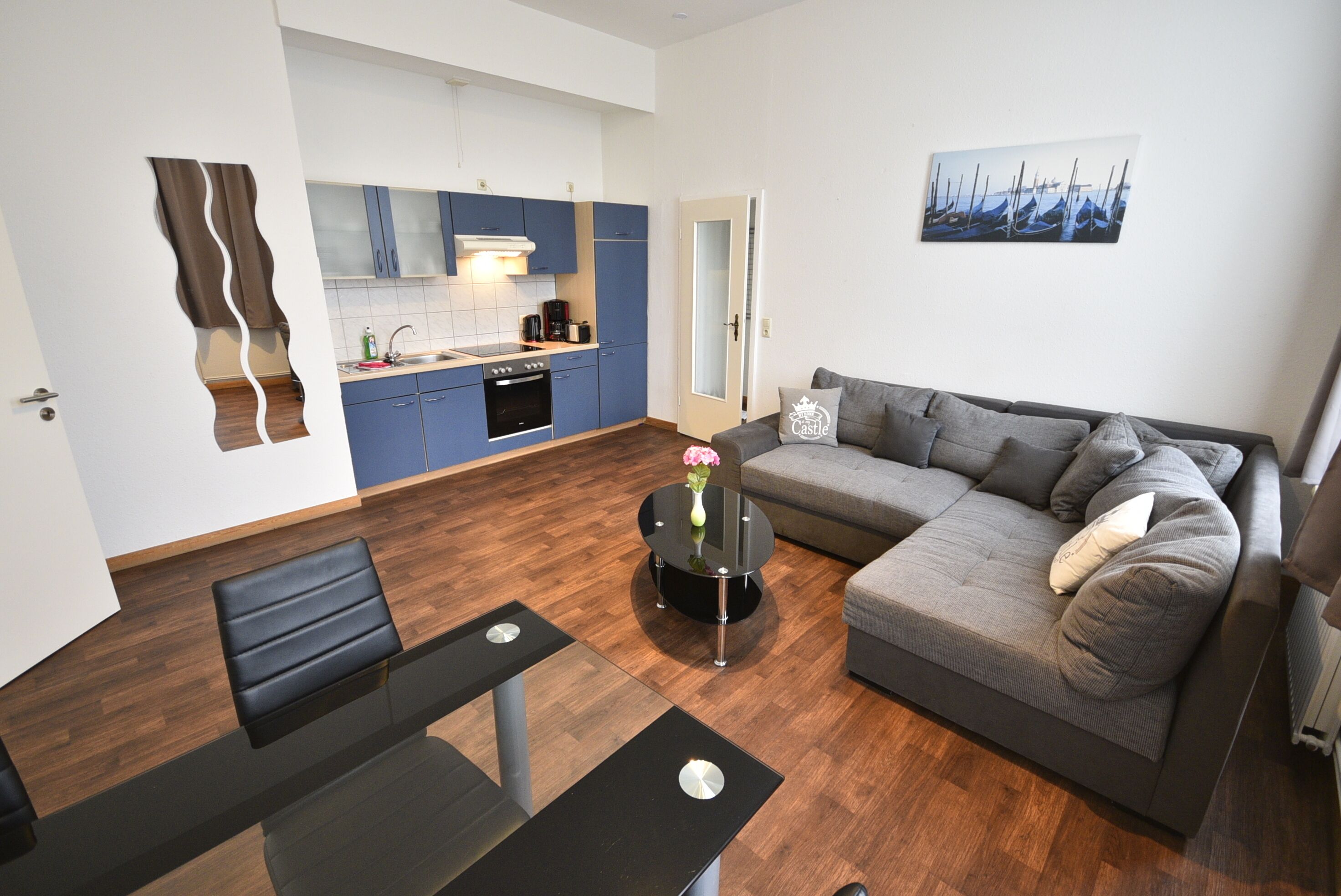 Una sala moderna con un sofá gris, una mesa de centro con un jarrón, y una cocina con armarios azules y una puerta blanca.