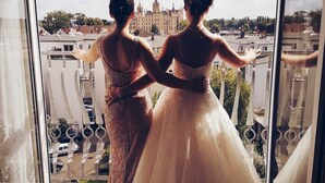 Dos mujeres con vestidos de novia de pie junto a un ventanal con vistas a un paisaje urbano.