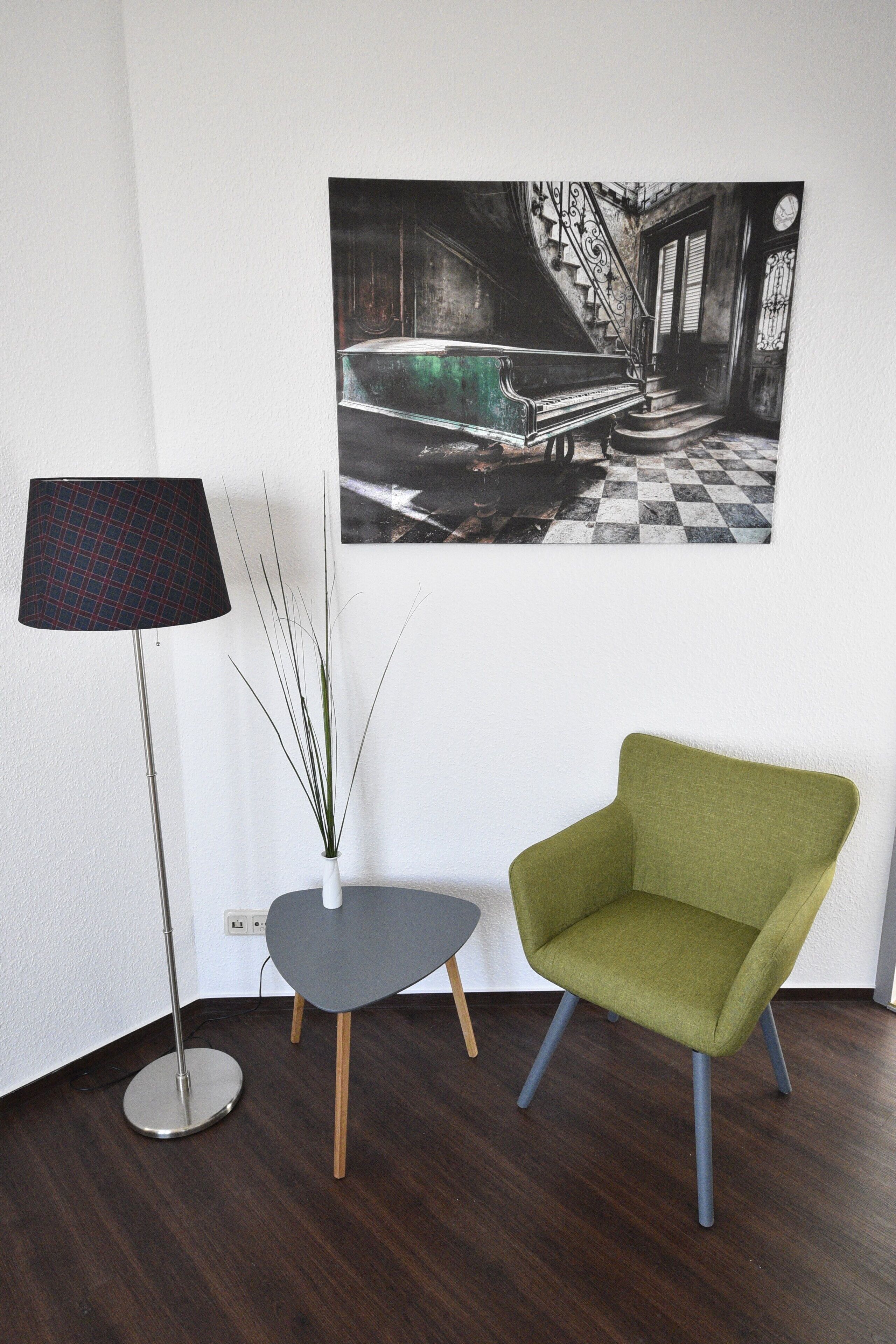 A room with a green armchair, a floor lamp, a small table, and a painting on the wall.