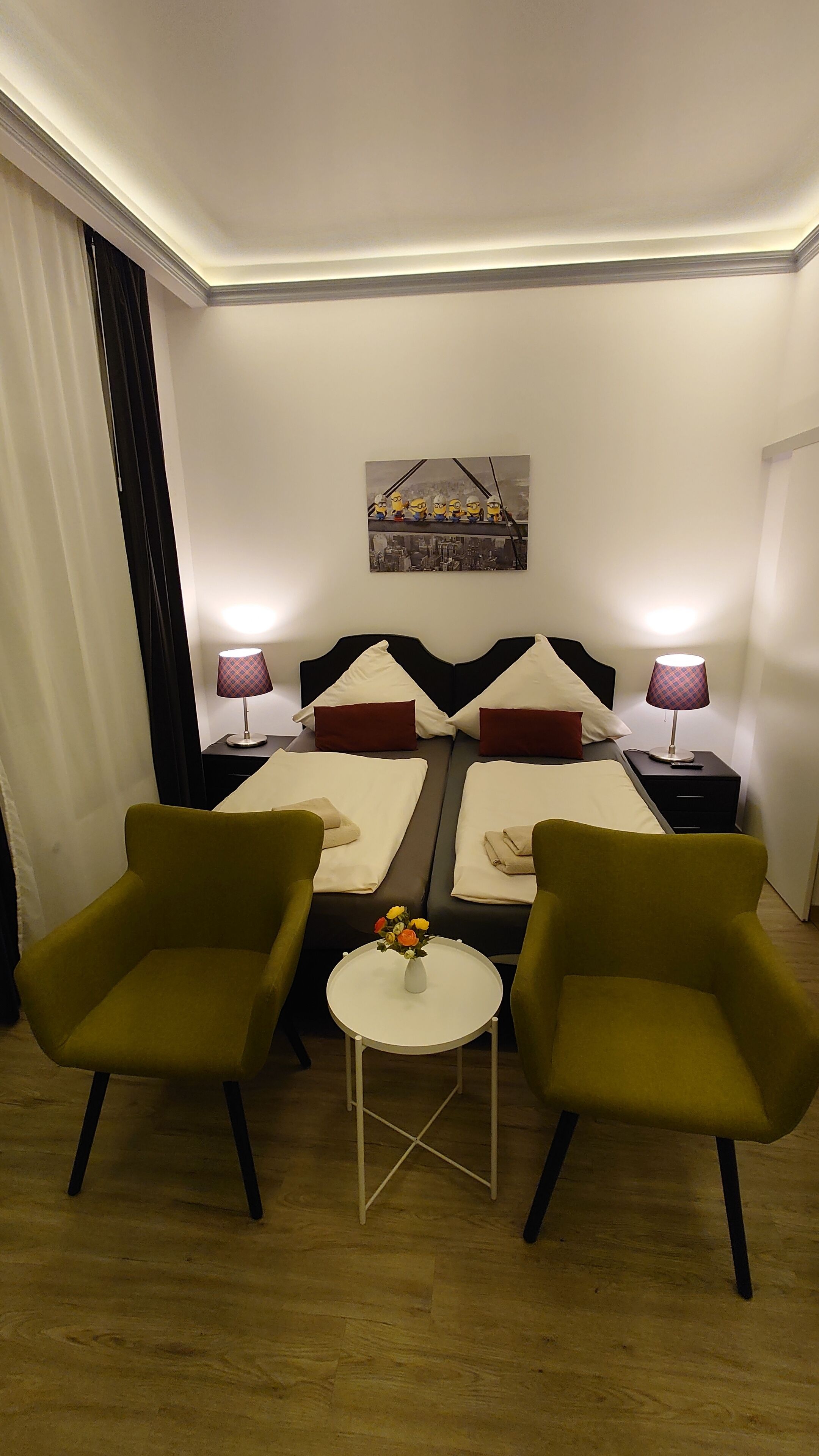 A hotel room with two beds, a small table with a vase of flowers, and two armchairs.