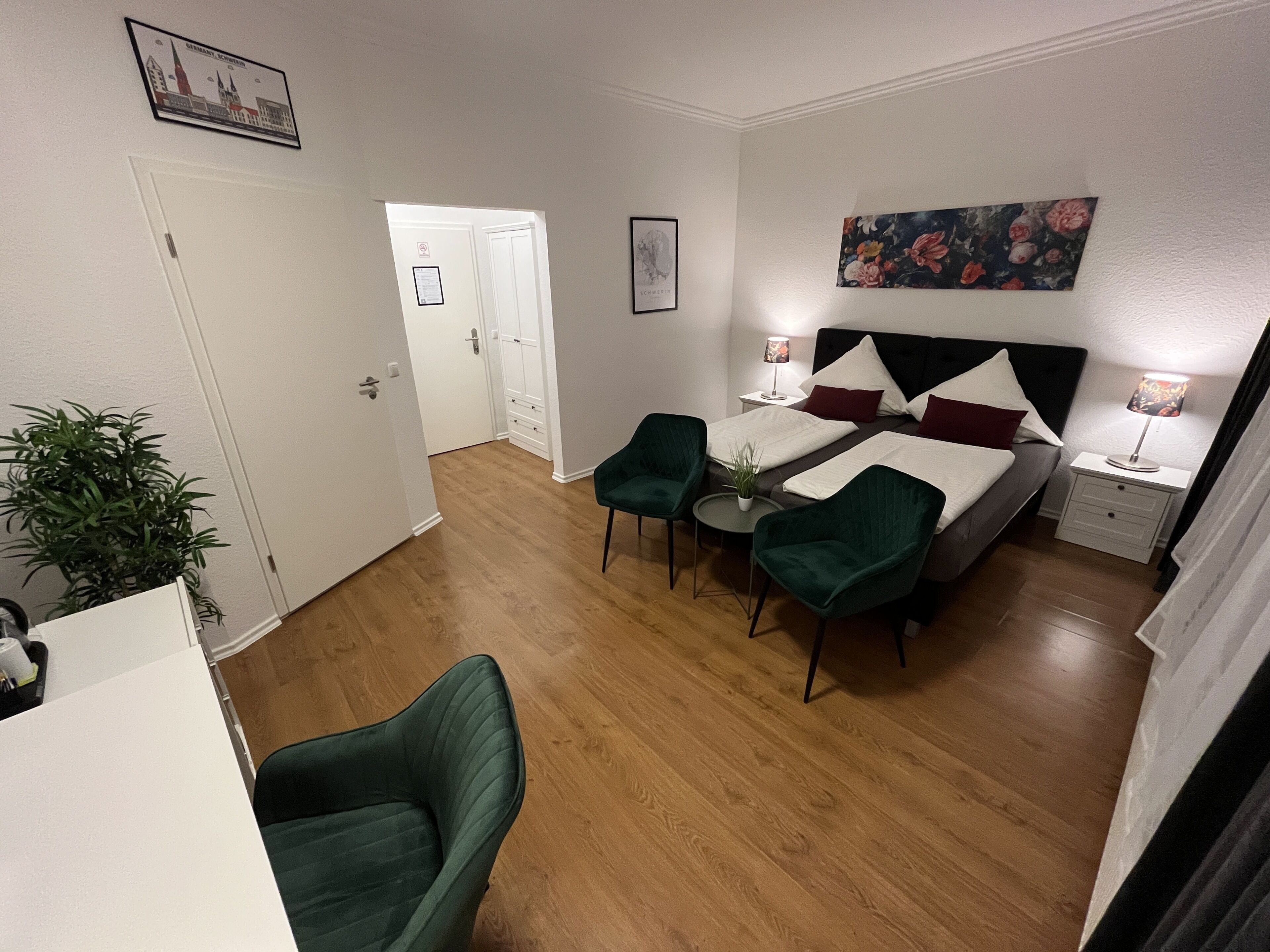 A hotel room with a bed, two green armchairs, a small table, a nightstand with a lamp, and a painting on the wall.