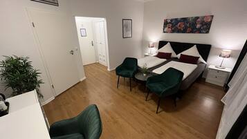 A hotel room with a bed, two green armchairs, a small table, a nightstand with a lamp, and a painting on the wall.
