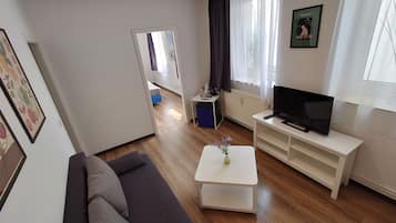 Una sala de estar con un sofá, un televisor sobre un mueble bajo, una mesa de centro con un florero y una cocina al fondo.