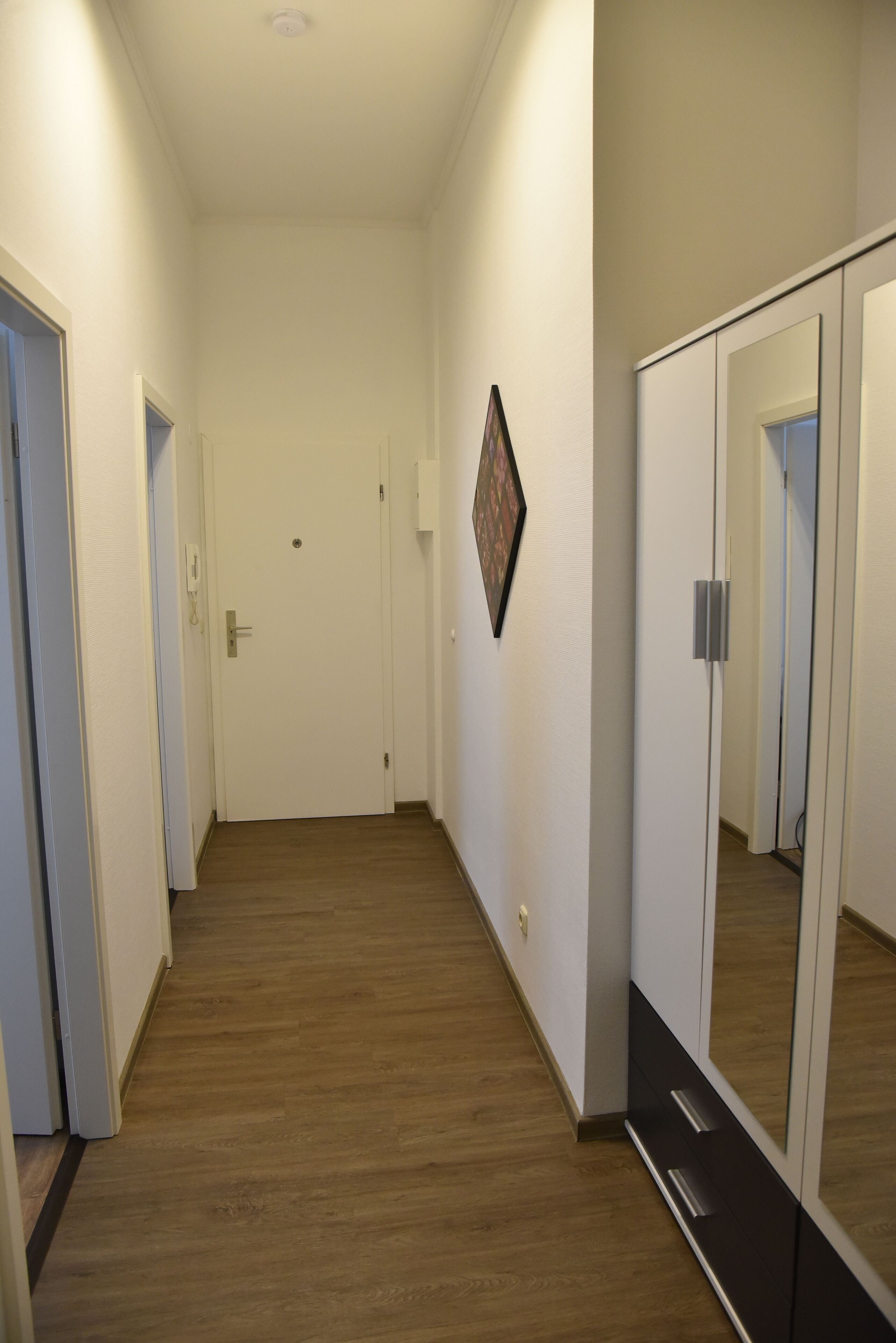 A hallway with wooden flooring, white walls, a door at the end, and a large mirror on the right.