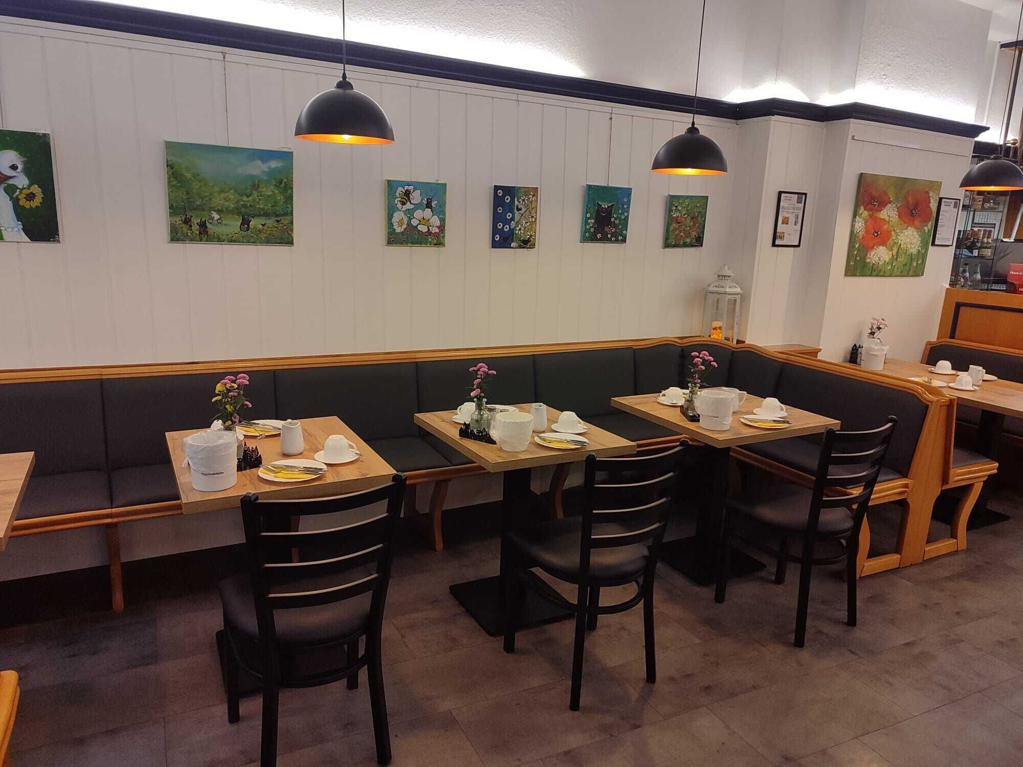 A restaurant interior with wooden tables and black cushioned booths, white paneled walls, and various paintings.
