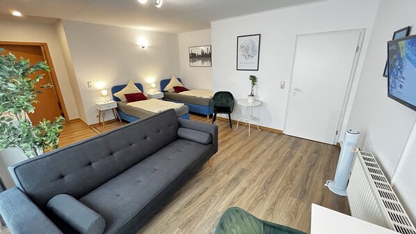 Una habitación moderna y luminosa con un sofá gris, una cama con ropa de cama azul y roja, una mesita redonda con una planta y una puerta blanca.