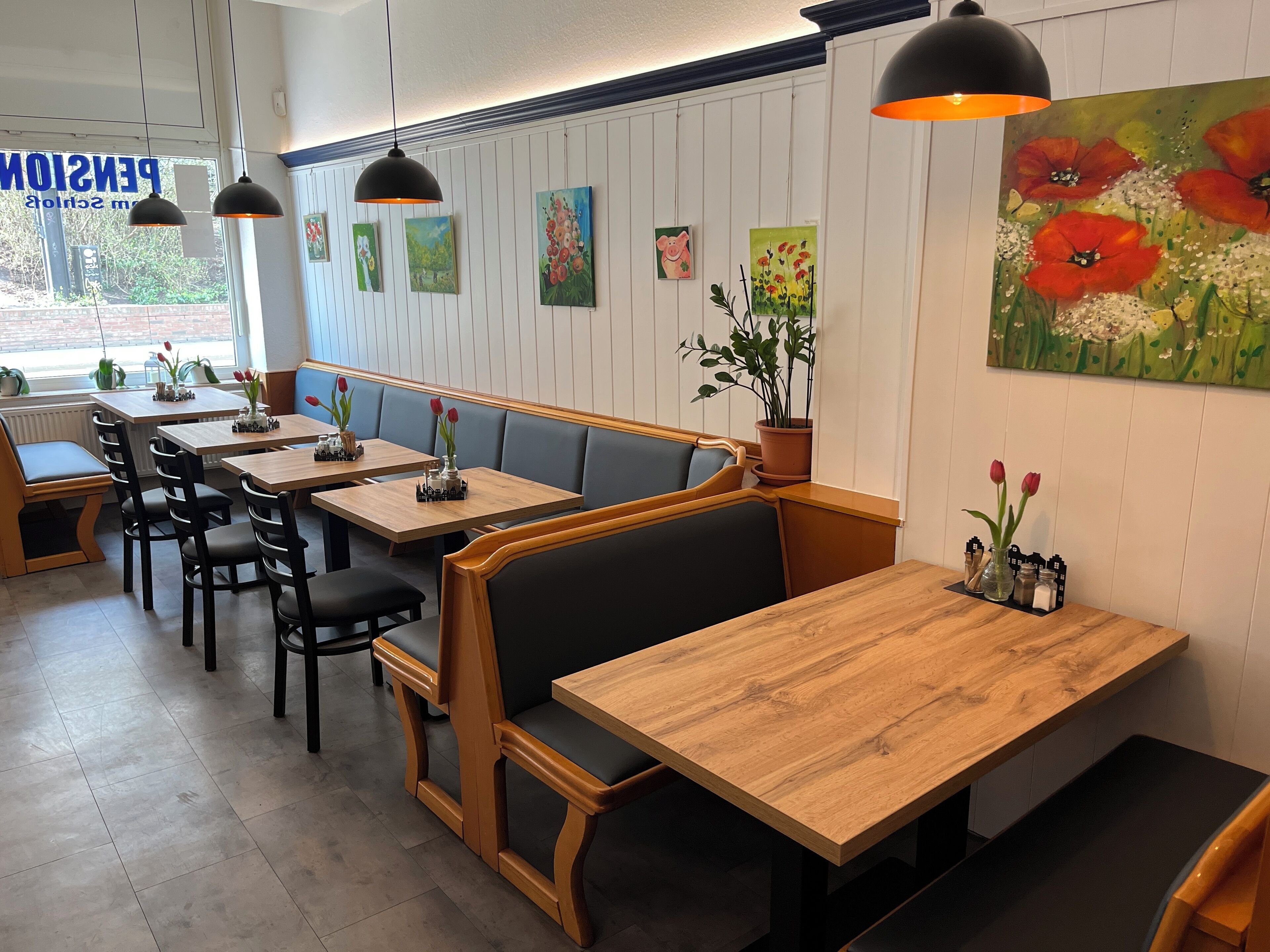 A restaurant interior with wooden tables and booths, potted plants, and artwork on the walls.