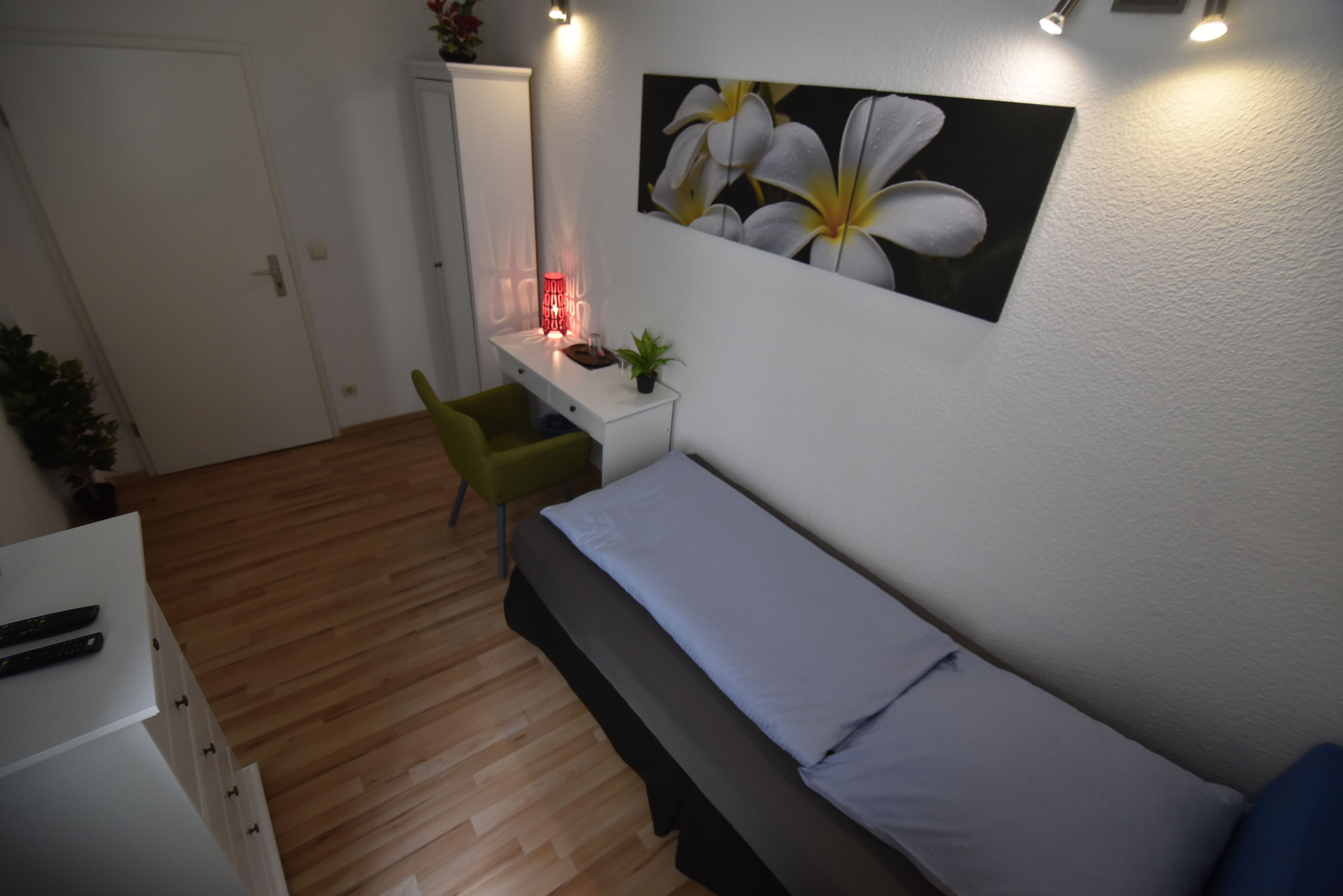 A bedroom with a bed, a desk, and a chair. There is a painting on the wall and a plant on the desk.