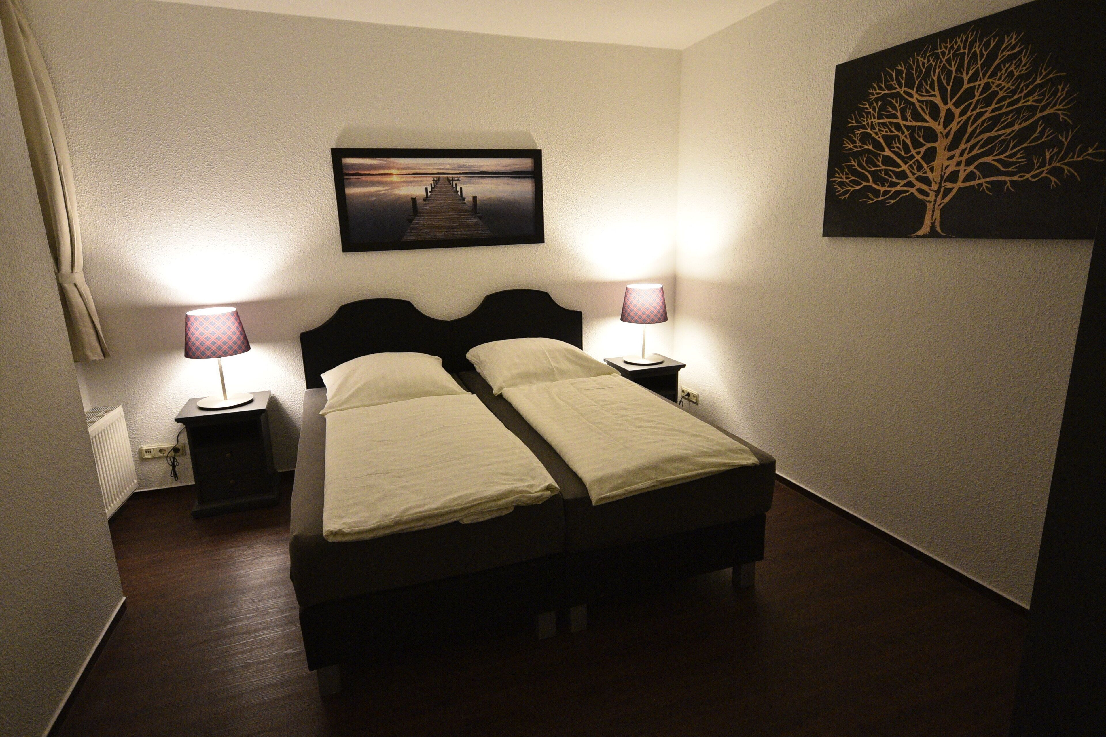 A double bed with white linens, two bedside tables with lamps, a framed picture of a tree, and a radiator on the wall.
