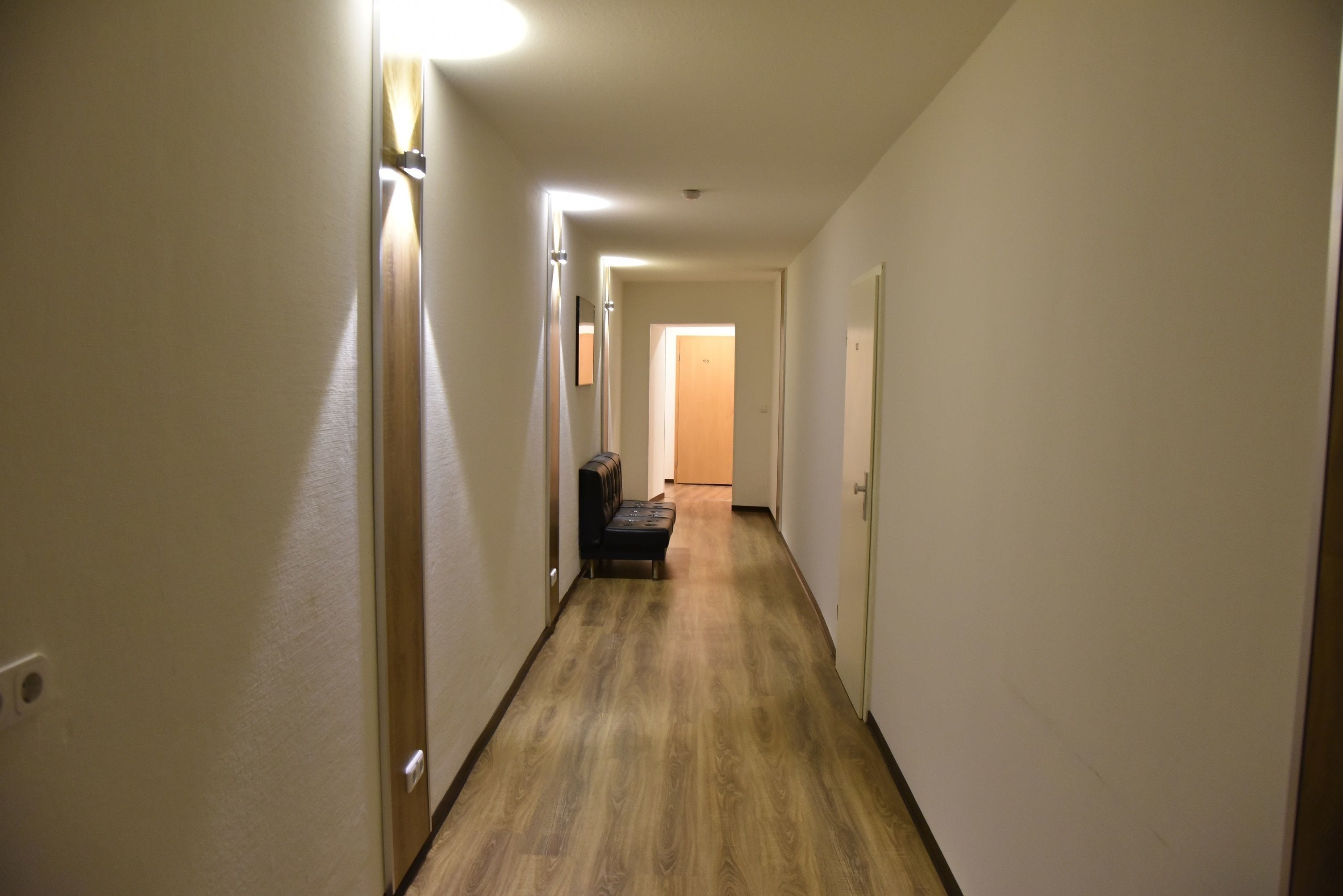 A narrow, well-lit hallway with wooden flooring, leading to a room with a bench and a door at the end.