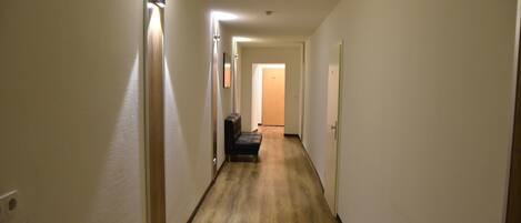 A narrow, well-lit hallway with wooden flooring, leading to a room with a bench and a door at the end.