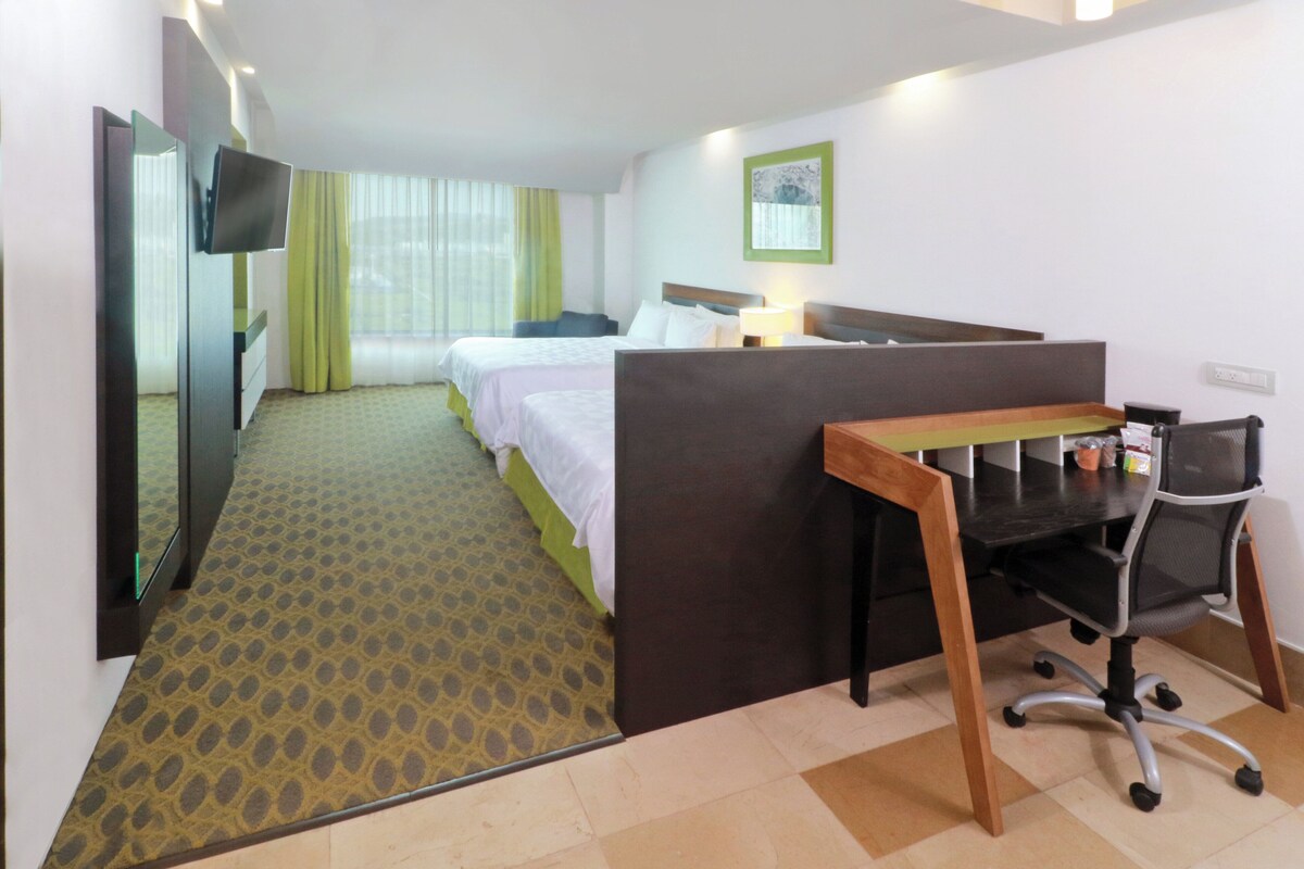 A hotel room with a bed, a desk with a chair, a TV, and a window with curtains.