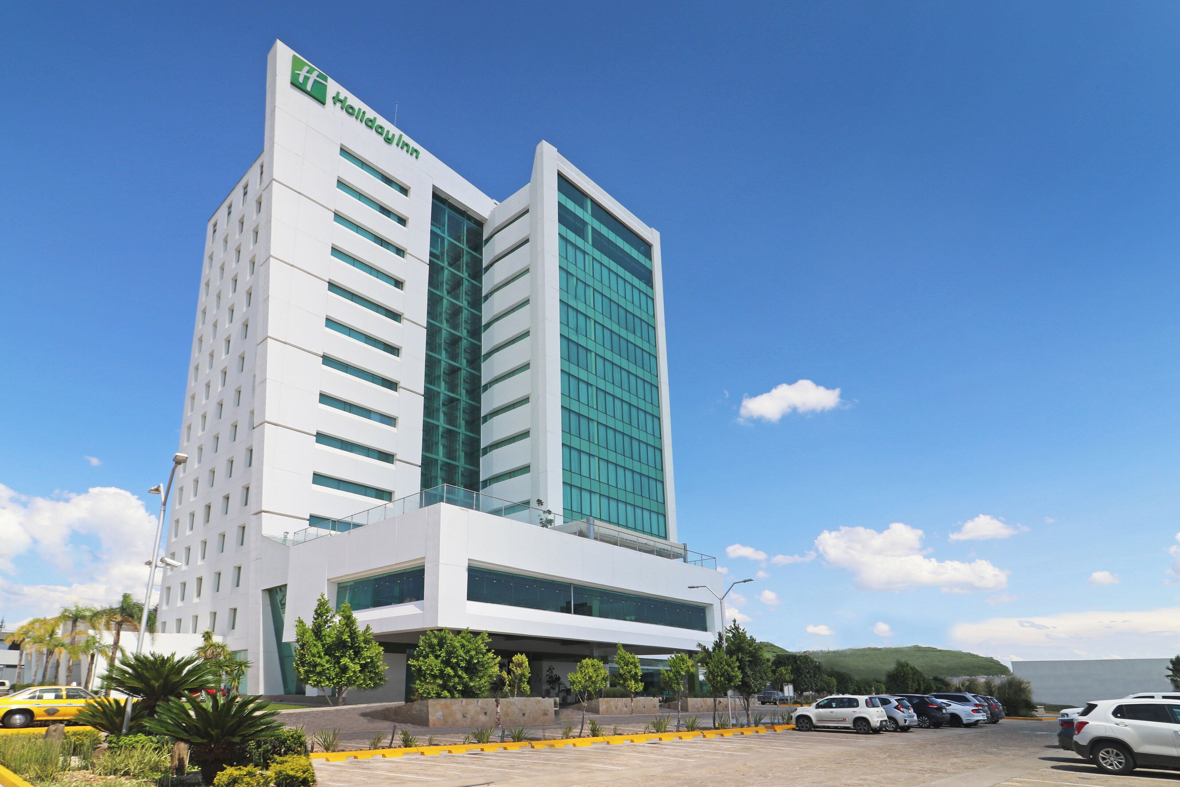 Un moderno hotel Holiday Inn con una fachada de vidrio destacada, un estacionamiento y un cielo azul despejado.