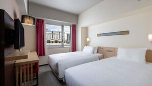 Habitación de hotel con dos camas, televisión, ventana con cortinas y vista a edificios.