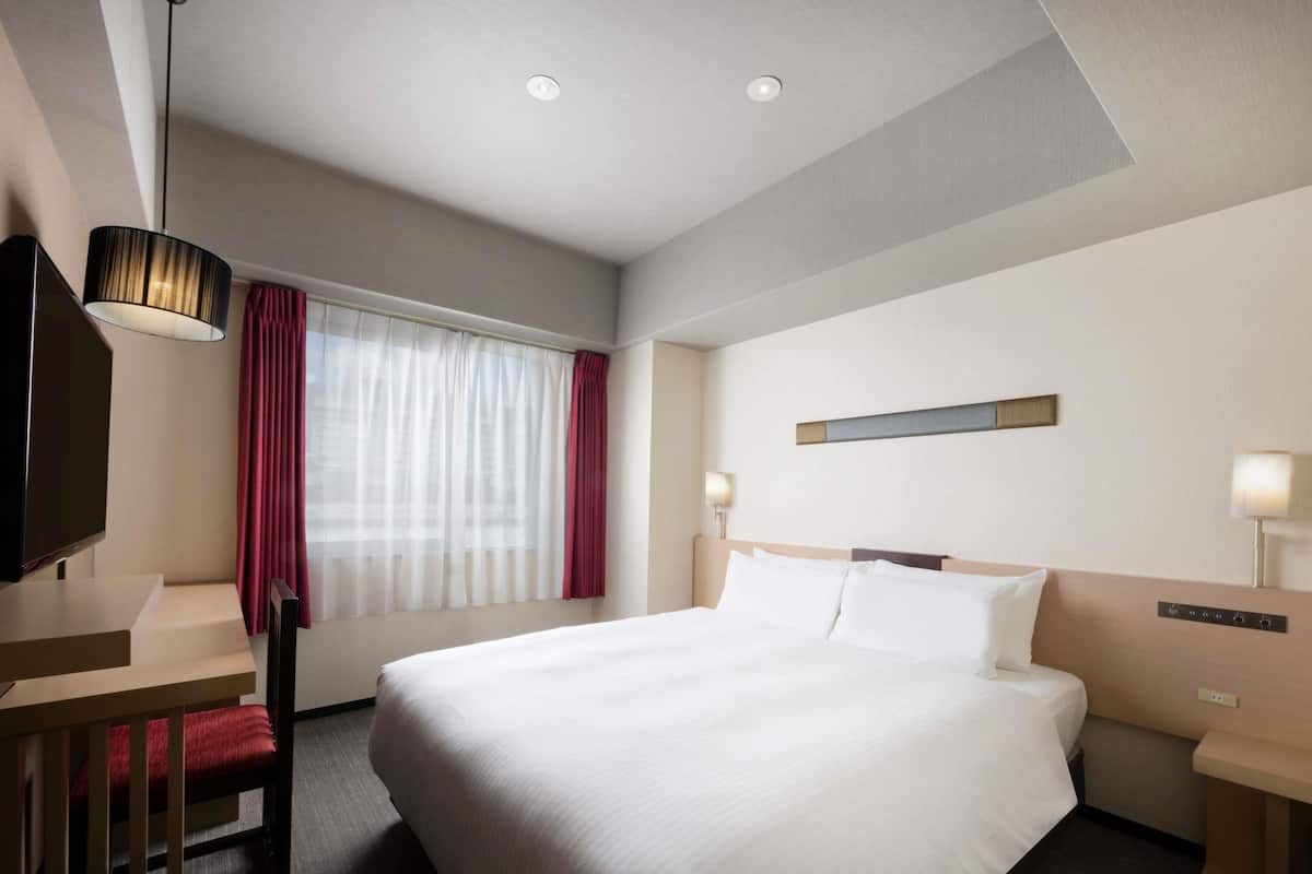 Habitació d'hotel amb un llit gran, una taula de treball, una cadira, un televisor i una finestra amb cortines.
