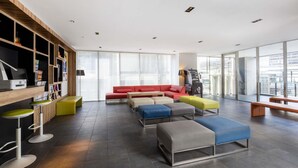 Un moderno hall de hotel con un sofá seccional rojo, otomanas coloridas, una zona de bar con taburetes, una máquina expendedora y grandes ventanales con vistas a la ciudad.