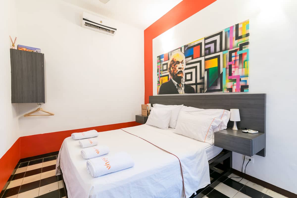 A hotel room with a bed, white linens, a nightstand, a lamp, and a colorful abstract painting on the wall.