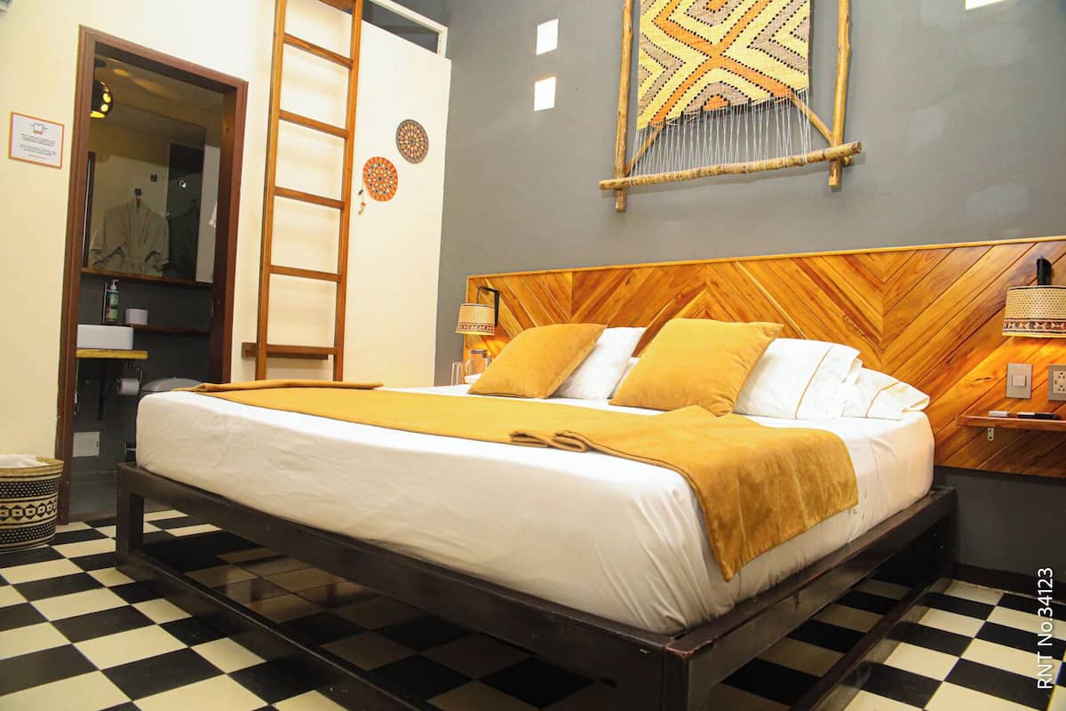 A hotel room with a checkered floor, a wooden bed frame, a large bed with white and yellow bedding, a wooden headboard, and a wall-mounted tapestry.