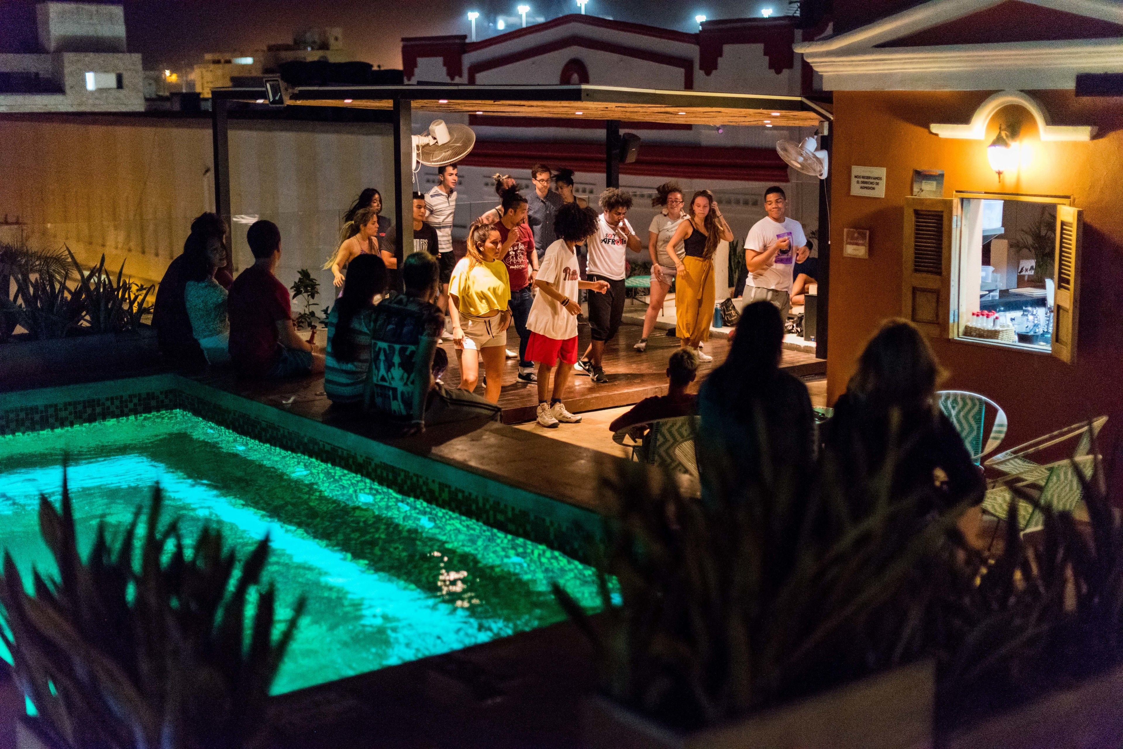 A group of people gathered around a pool with a fountain, surrounded by a building with a balcony and a sign indicating a restaurant.
