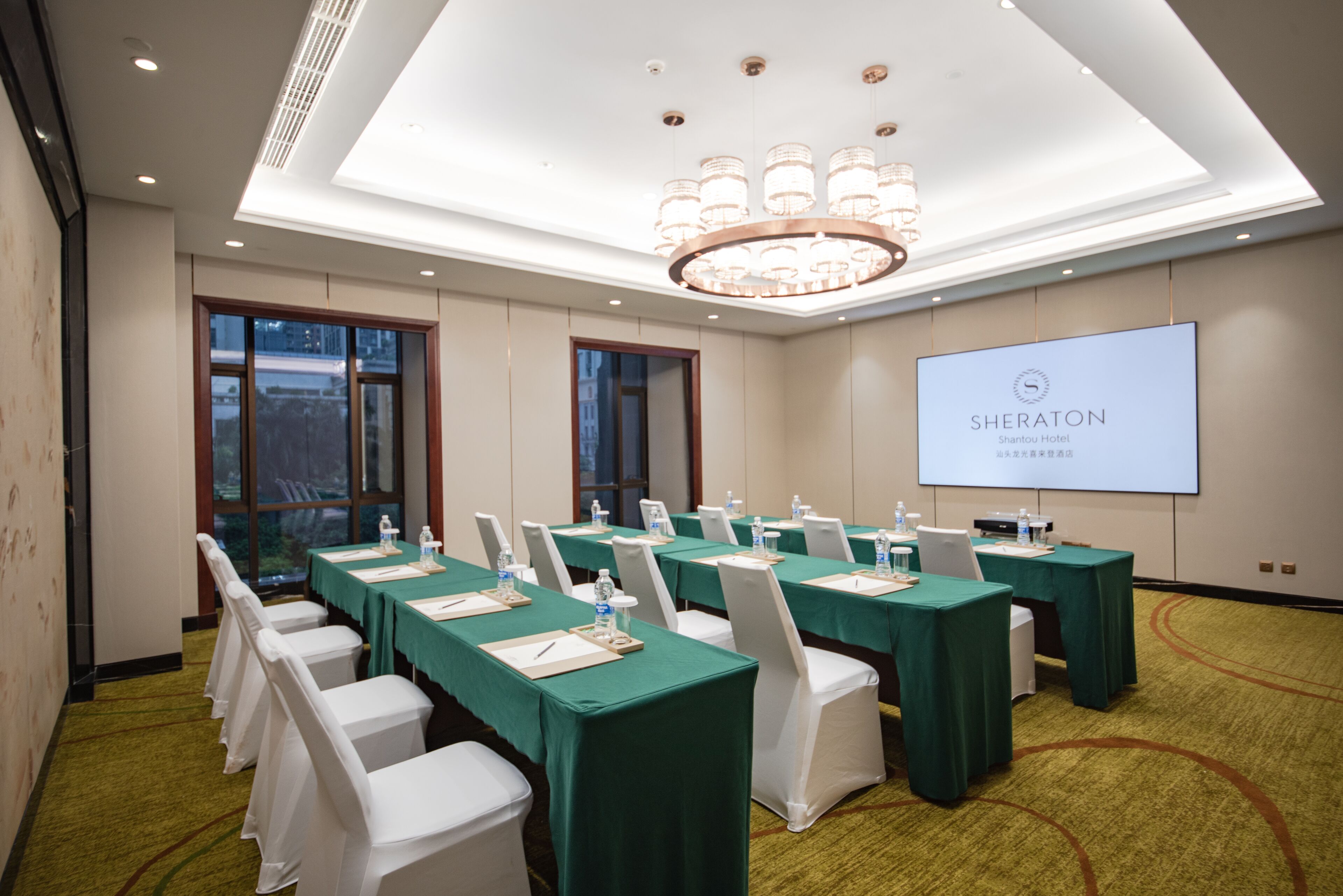 A conference room with green tables and white chairs, a large screen displaying 'SHERATON Shantou Hotel', and a chandelier.