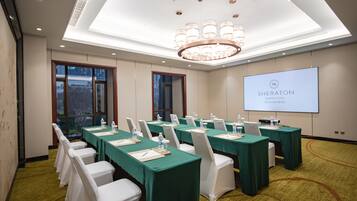 Un salón de conferencias con mesas verdes y sillas blancas, una pantalla grande mostrando 'SHERATON Shantou Hotel' y un candelabro.