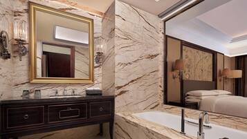 Un baño lujoso con tocador de mármol, un espejo grande, una bañera y una cama que se ve en el reflejo.