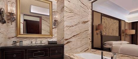 A luxurious bathroom with a marble vanity, a large mirror, a bathtub, and a bed visible in the reflection.
