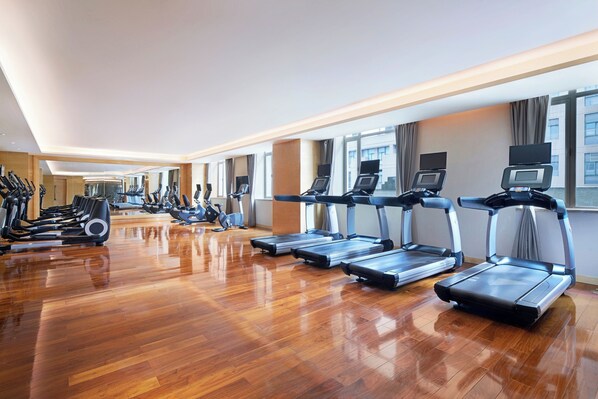 A spacious gym with multiple treadmills and exercise bikes, large windows, and a polished wooden floor.