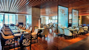 Un restaurante moderno con listones de madera en el techo, un área central de mesas con sillas acolchadas y una zona de bar con taburetes altos.