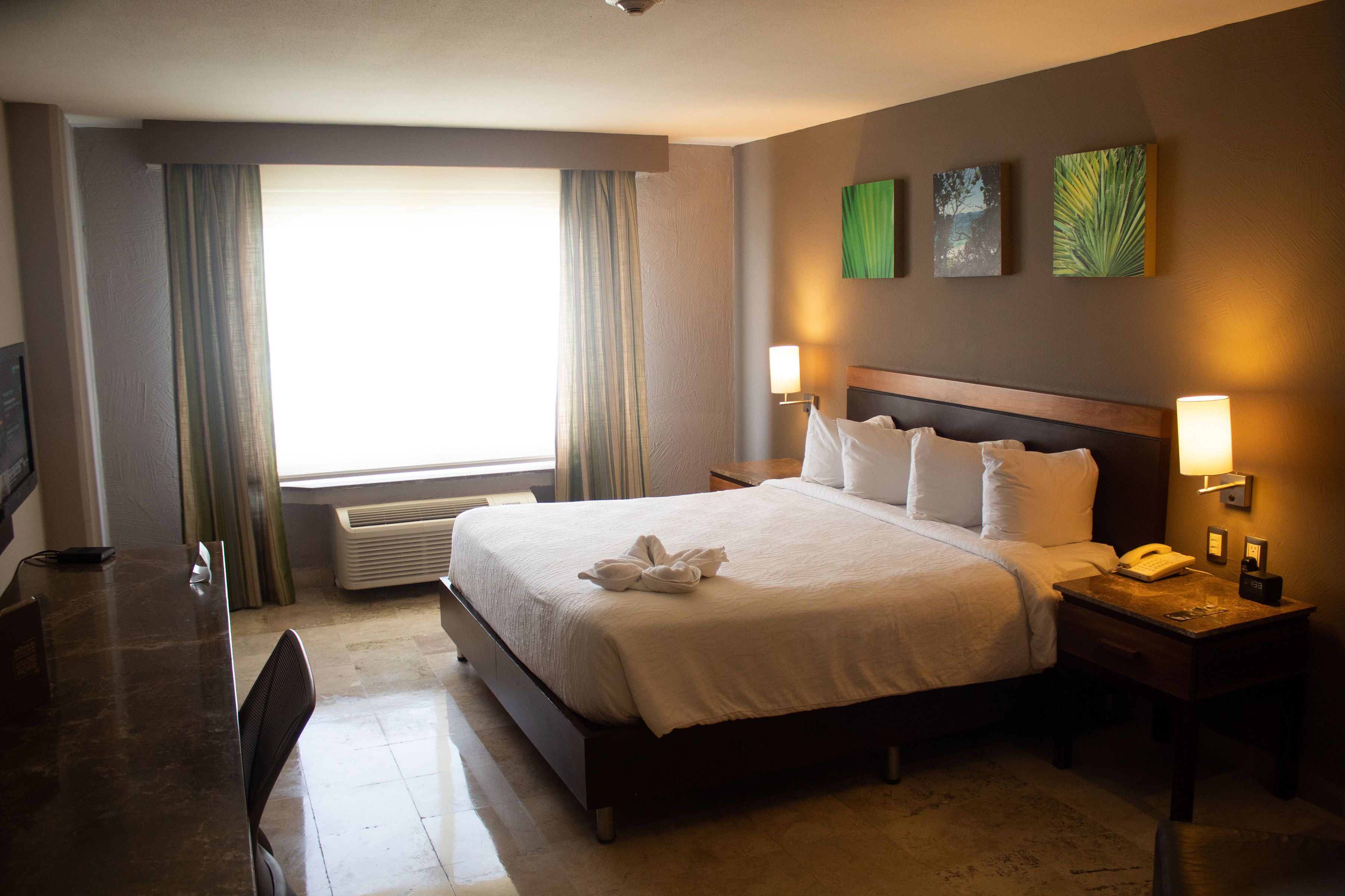 A hotel room with a large bed, bedside lamps, a desk with a chair, and a window with curtains.