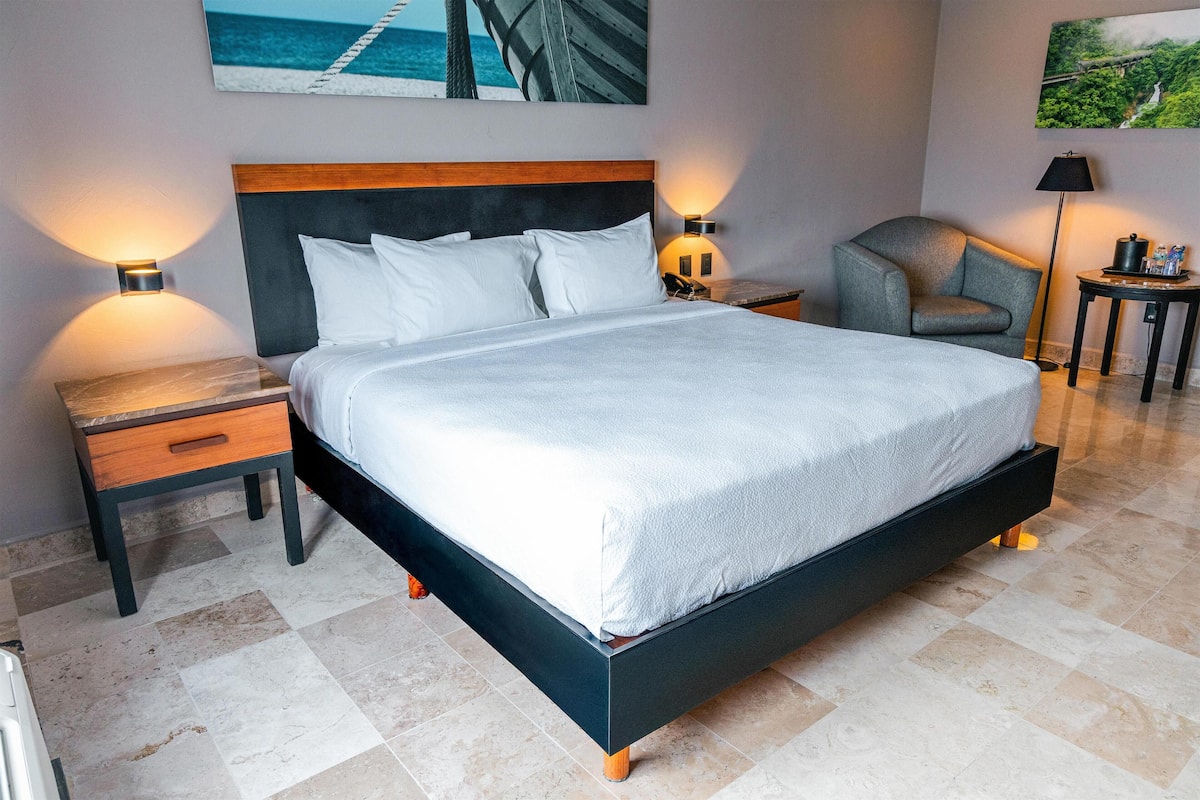 A modern hotel room with a large bed, bedside tables, a chair, and artwork on the walls.