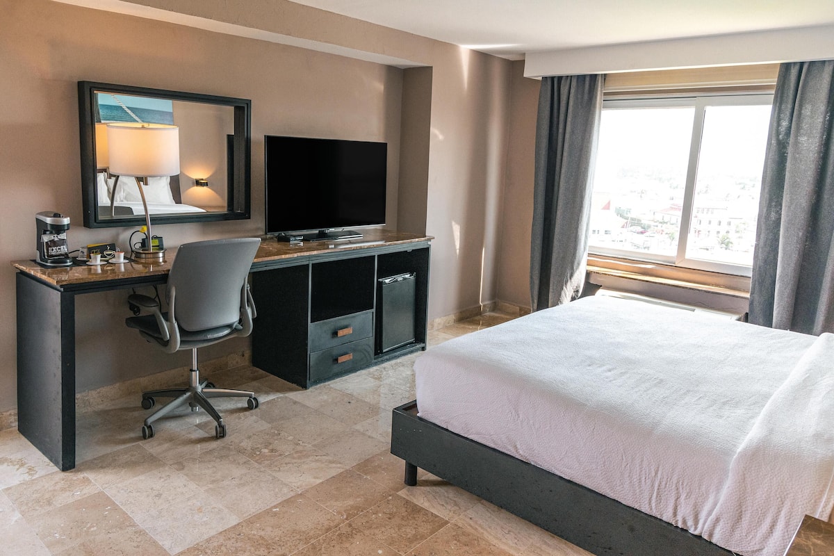 A hotel room with a bed, a desk with a computer, a chair, a television, and a window with curtains.