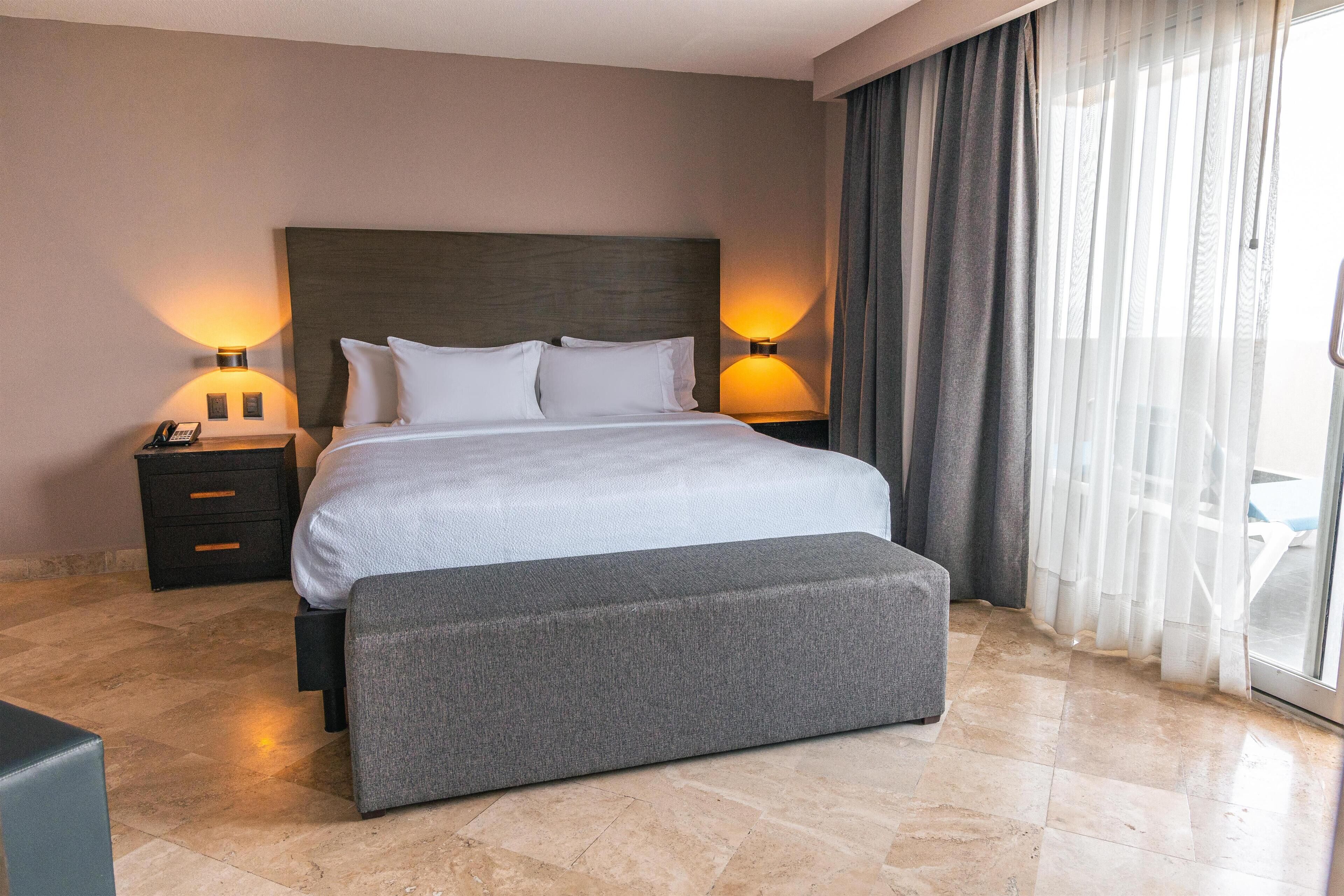 Une chambre d’hôtel moderne avec un grand lit, deux tables de chevet avec des lampes, un banc et une vue dégagée à travers des rideaux légers.