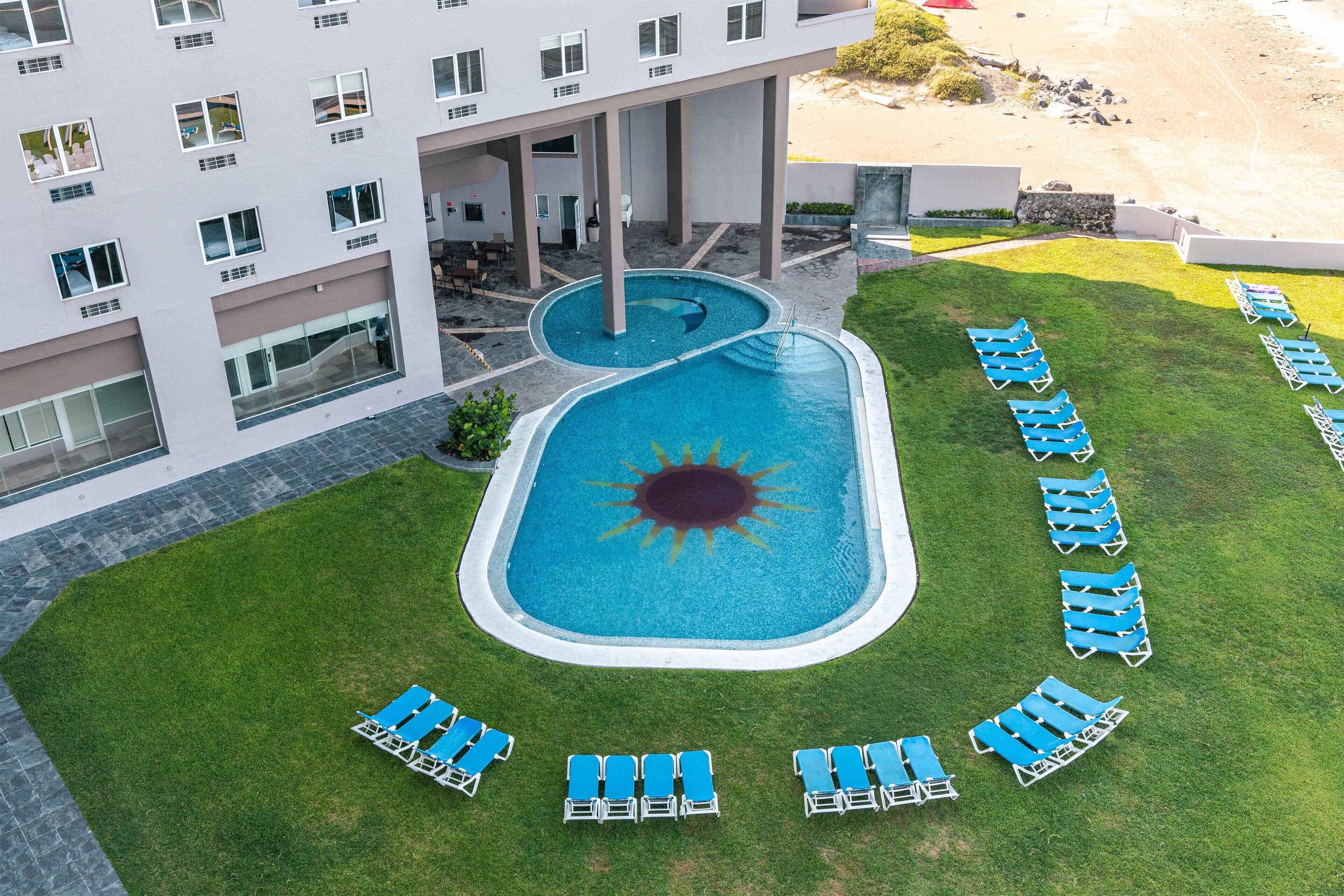 A rooftop pool with a sun design, surrounded by blue lounge chairs, and a building with multiple windows and balconies.