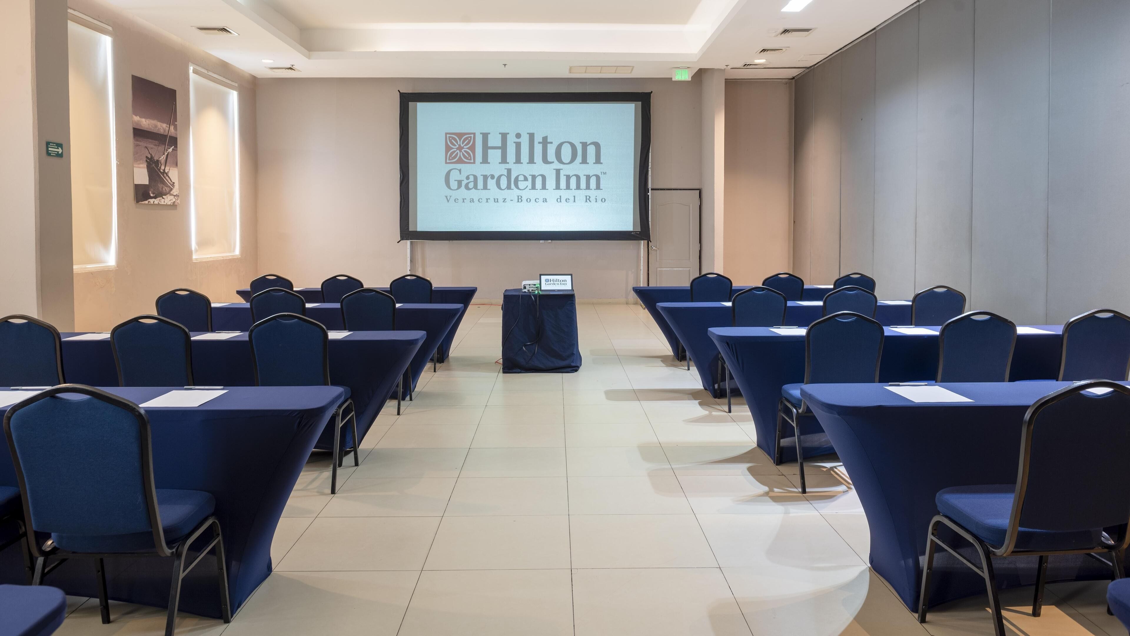 Un salón de conferencias con filas de sillas y mesas azules, un atril y una pantalla que muestra el logotipo de Hilton Garden Inn.