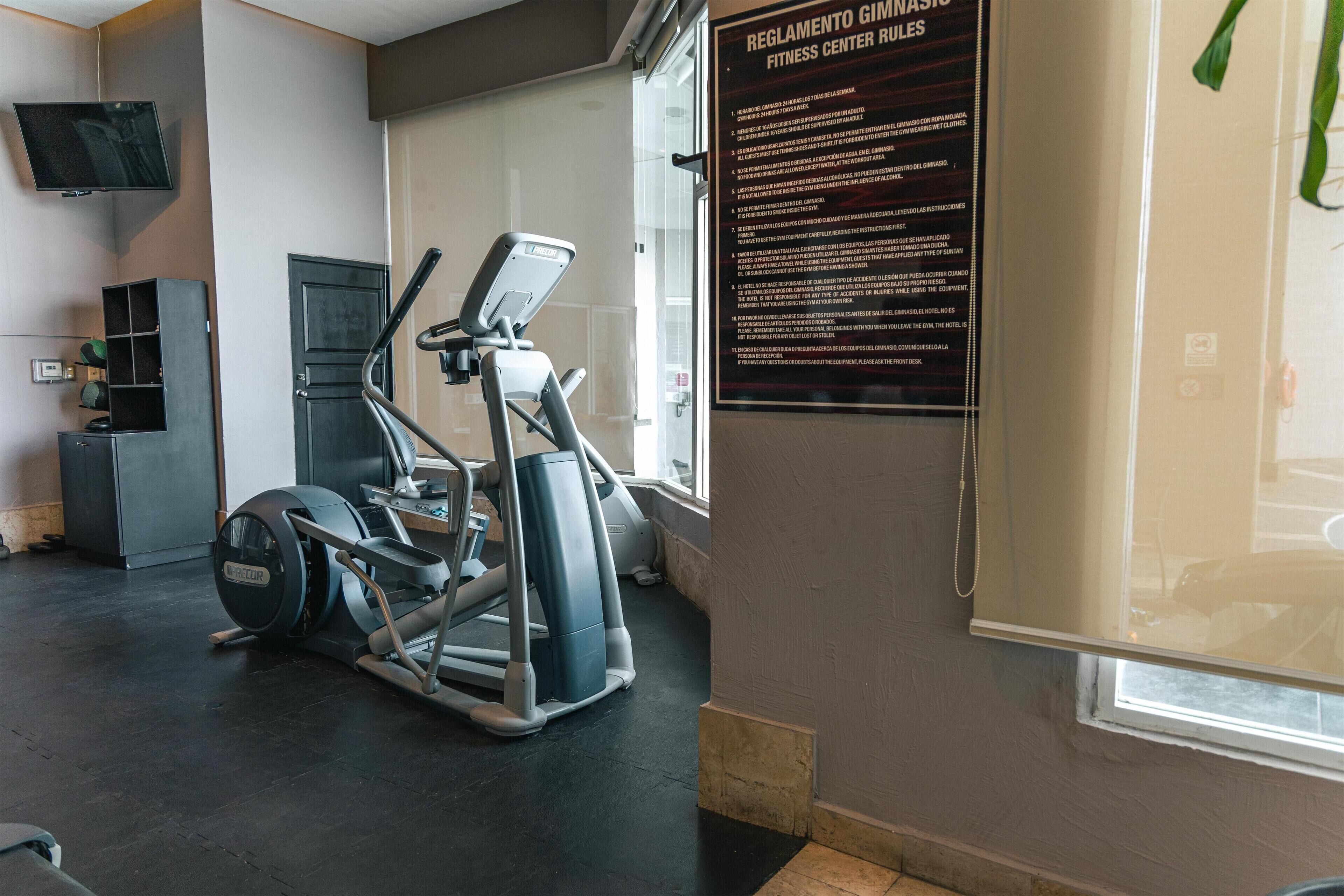 Gimnasio con equipo de ejercicio, un televisor y un cartel con normas.