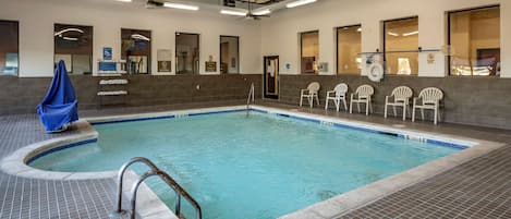 An indoor swimming pool with tiled flooring, a diving board, and several white chairs along the poolside.