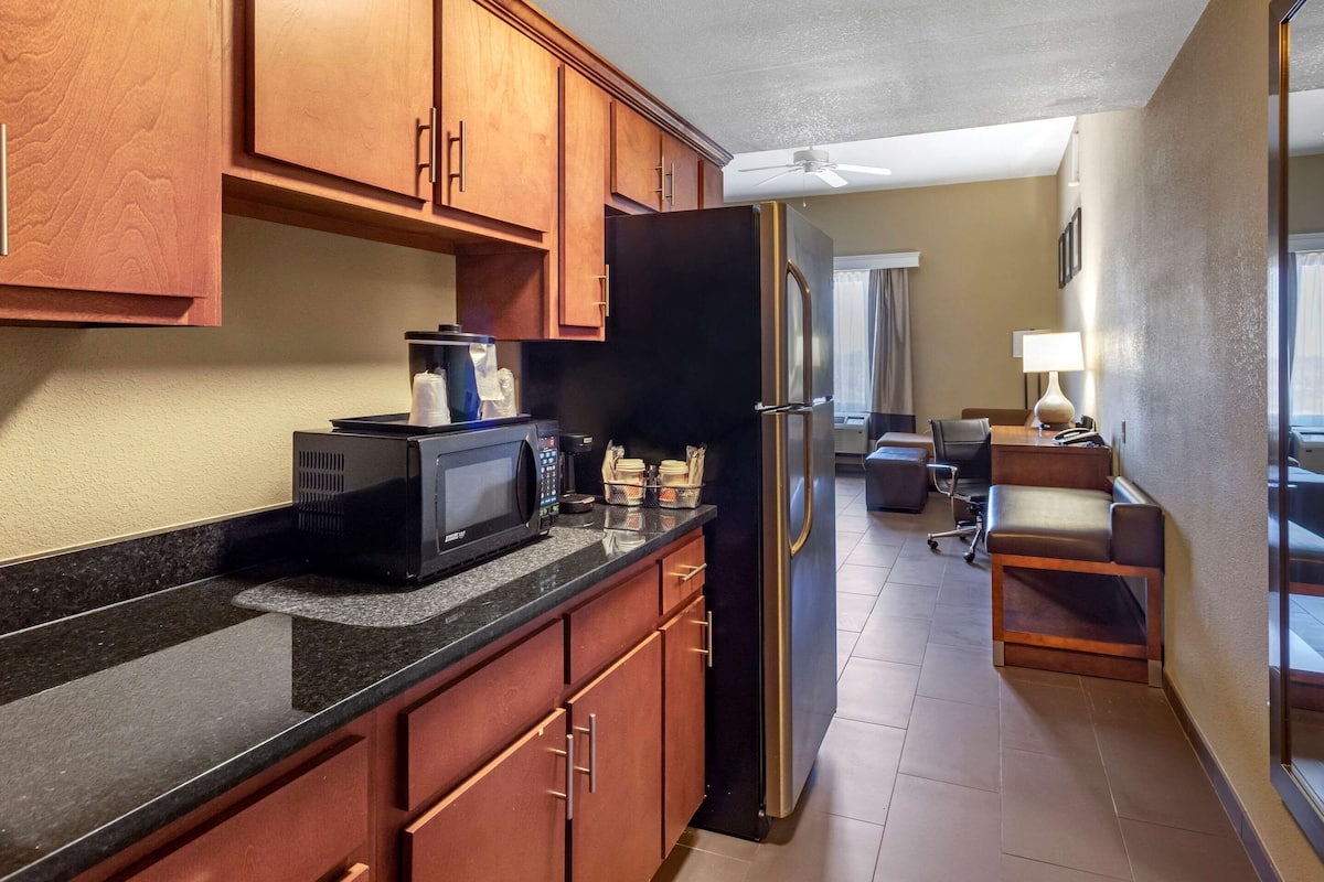 A modern kitchen with wooden cabinets, a black refrigerator, and a microwave. There is a dining area with a table and chairs, and a window with curtains.