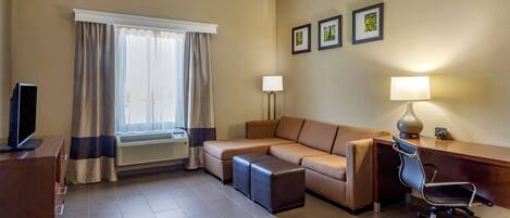 A hotel room with a sofa, ottoman, desk, chair, and a television.