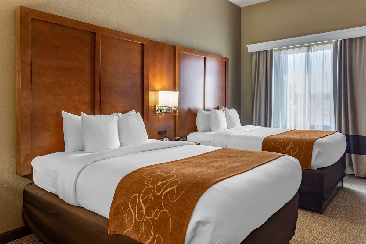 A hotel room with two beds, a wooden headboard, a nightstand with a lamp, and a window with curtains.