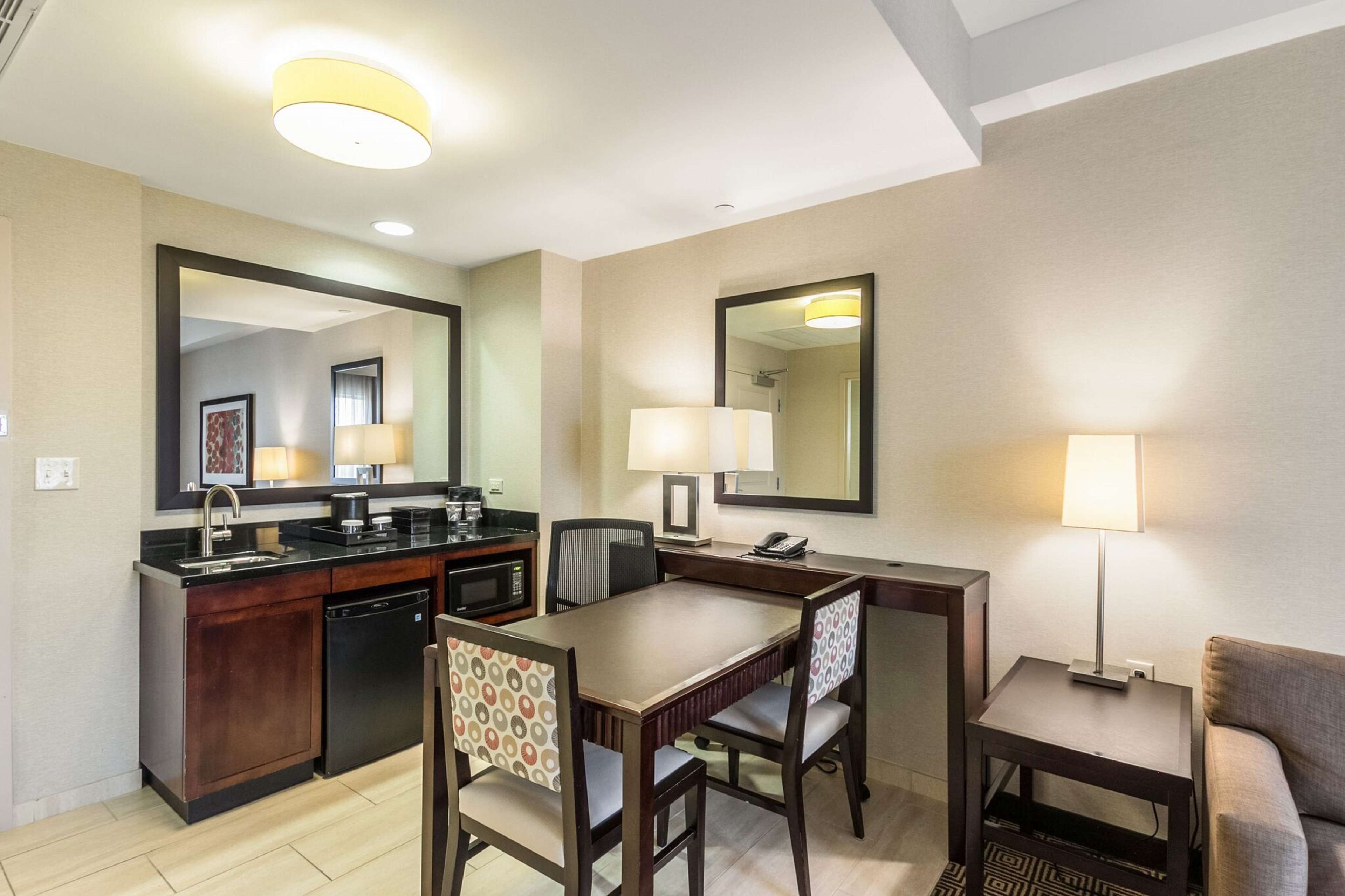A compact hotel room with a kitchenette, a dining area with a table and chairs, a sofa, and a large mirror.