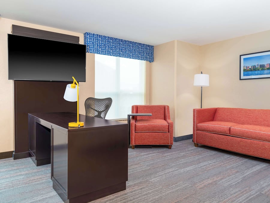 Une chambre d’hôtel avec un bureau en bois foncé, un canapé rouge, un fauteuil rouge, une grande fenêtre agrémentée d’un lambrequin bleu, un téléviseur à écran plat, une lampe sur pied et un tableau encadré au mur.