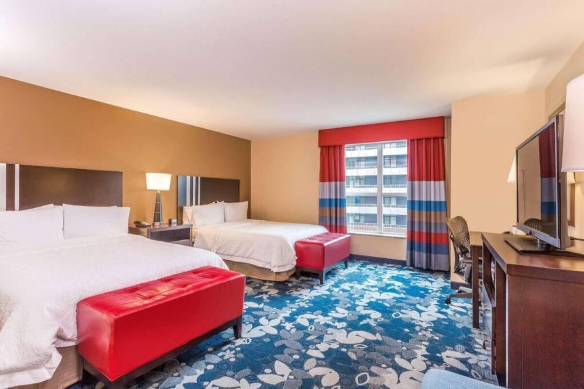 Quarto de hotel com duas camas, uma mesa, uma televisão e uma janela com cortinas.