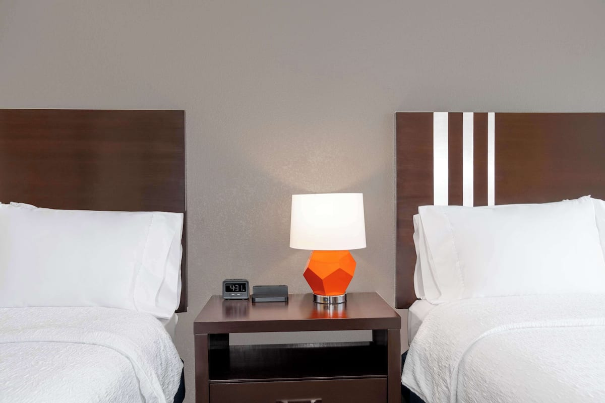 Una habitación de hotel con dos camas, una mesita de noche con lámpara y un reloj digital.